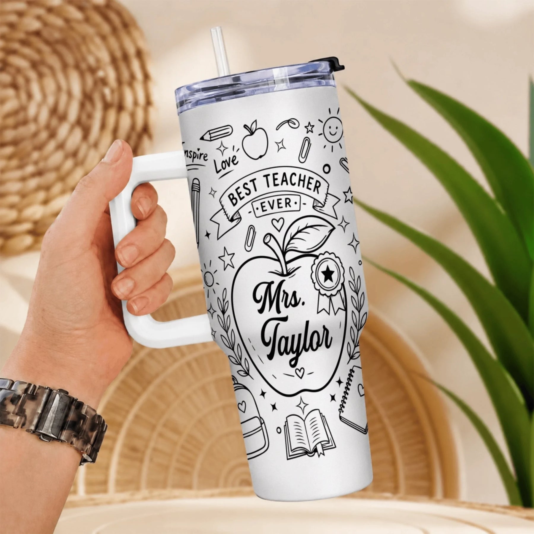 A hand holds a white insulated tumbler with a handle and straw, decorated with black line art of school items and the text "Best Teacher Ever" and "Mrs. Taylor".