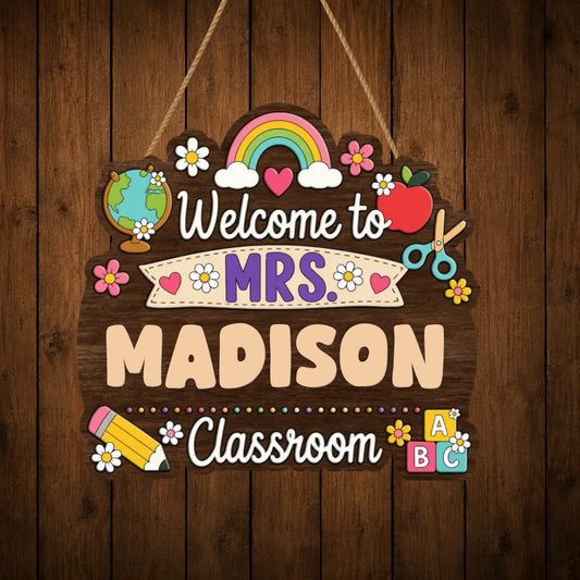 A scalloped wooden sign with "Welcome to MRS. MADISON Classroom" text, decorated with a rainbow, apple, globe, scissors, and flowers, against a wood background.