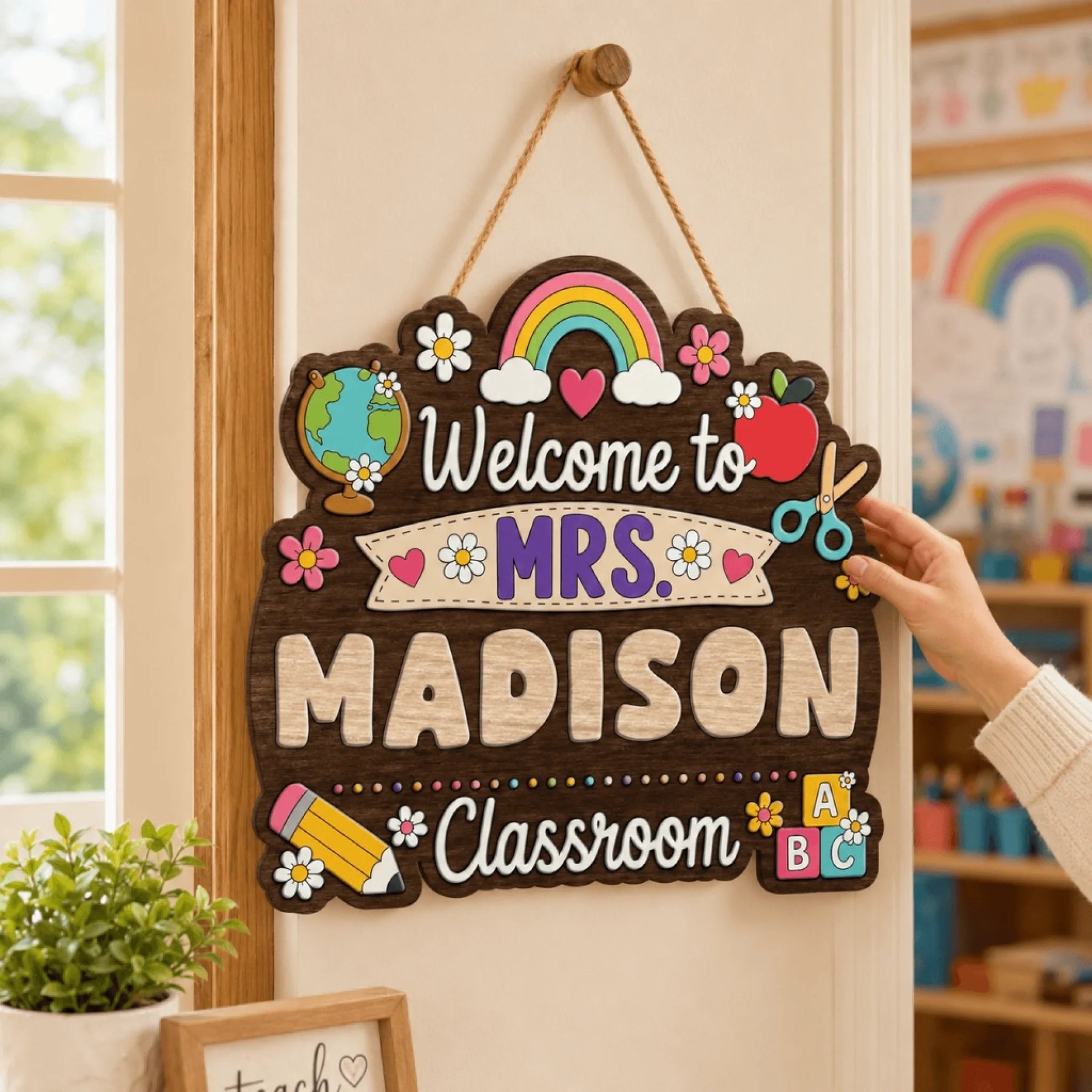 A hand gently touches a colorful "Welcome to MRS. MADISON Classroom" wooden sign hanging on a wall next to a window in a bright classroom.