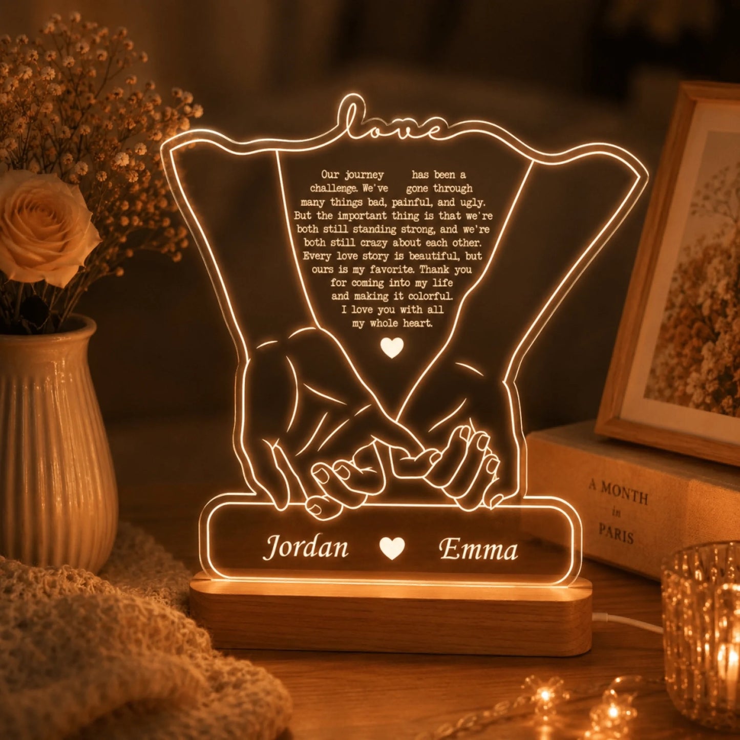 An illuminated lamp with two hands holding, a romantic quote, and names "Jordan" and "Emma" on a wooden base, surrounded by a rose and books.