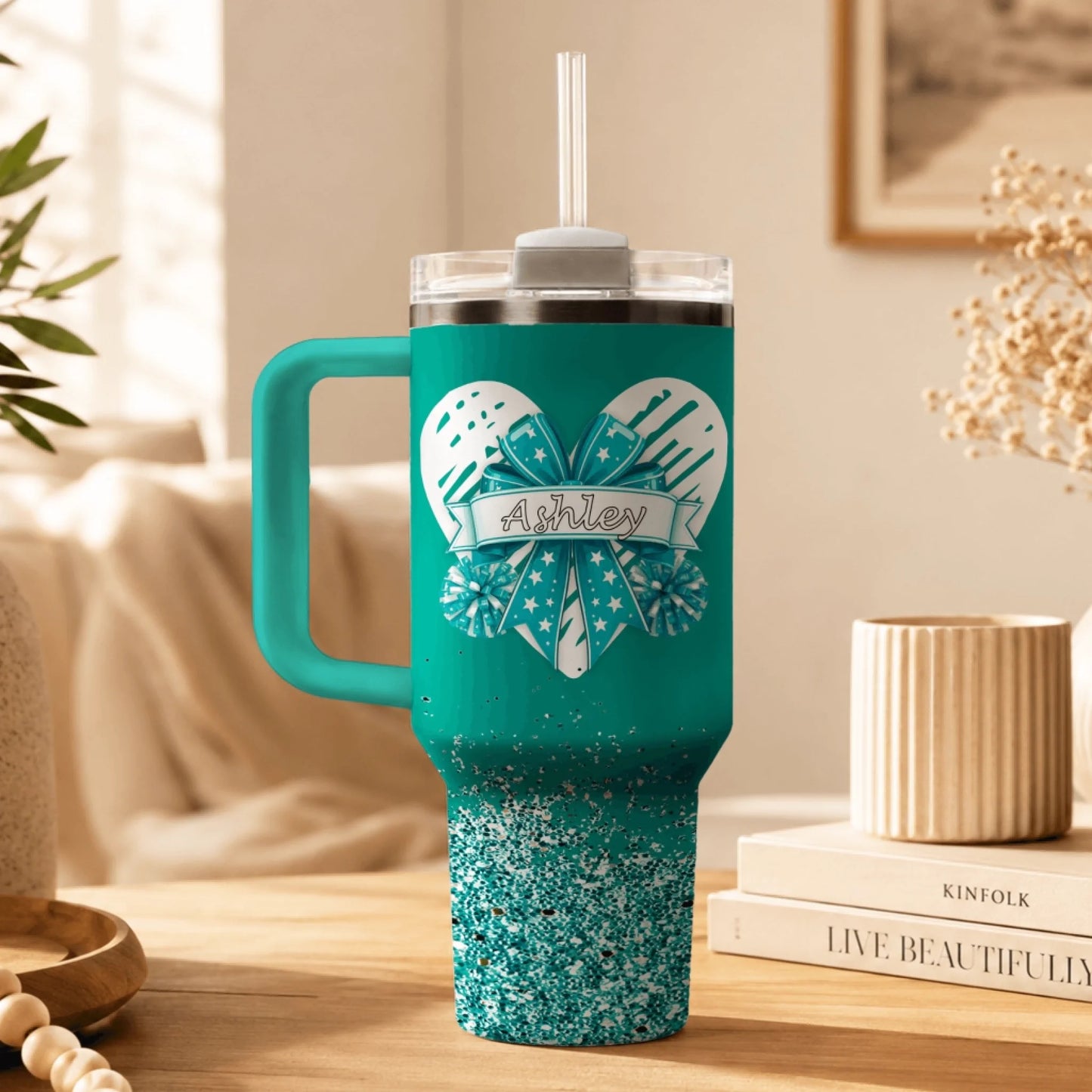 A personalized teal tumbler for cheerleaders decorated with a heart bow design and the name Ashley printed in black.