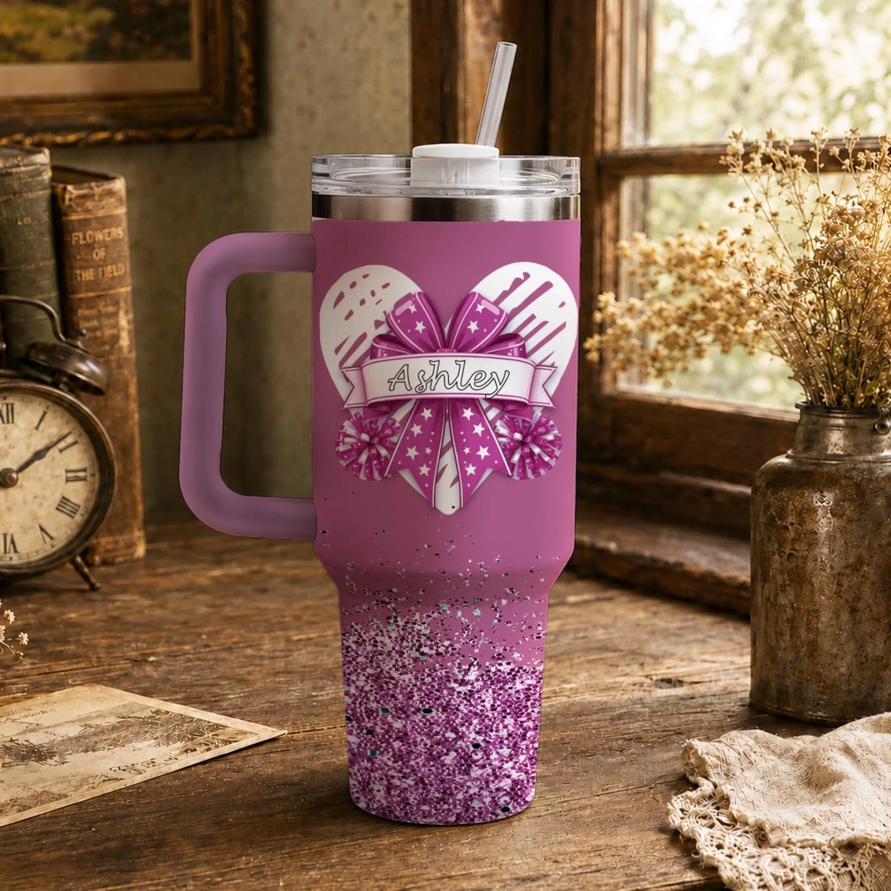 A personalized pink tumbler with a cheer themed heart and bow, featuring the name Ashley on a wooden desk.