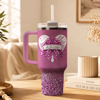 A custom pink insulated tumbler featuring a heart, bow, and pom poms design with the personalized name Ashley in script.