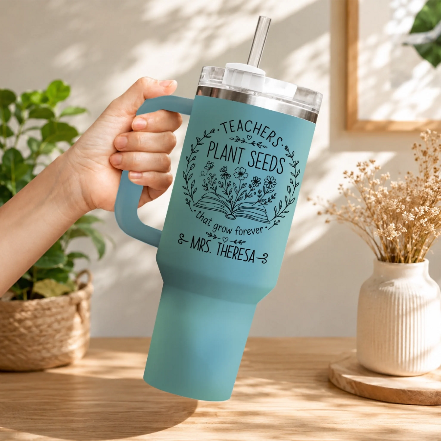 Hand holding a blue personalized teacher tumbler with floral and book graphic and straw lid.