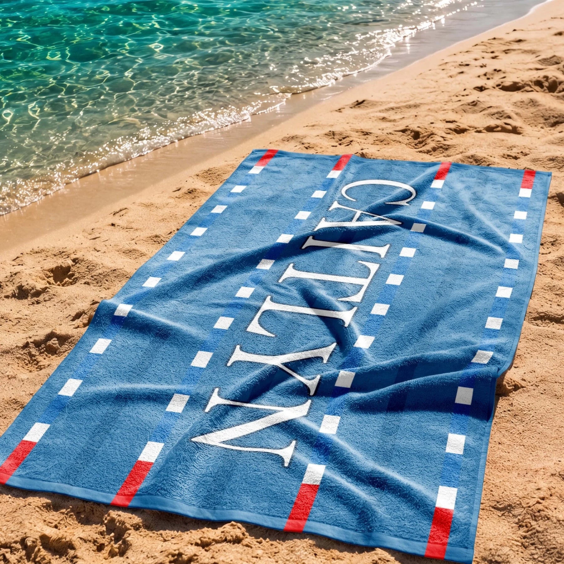Personalized blue swimming pool lane beach towel with the name Caitlyn, displayed on a sandy beach by the water.