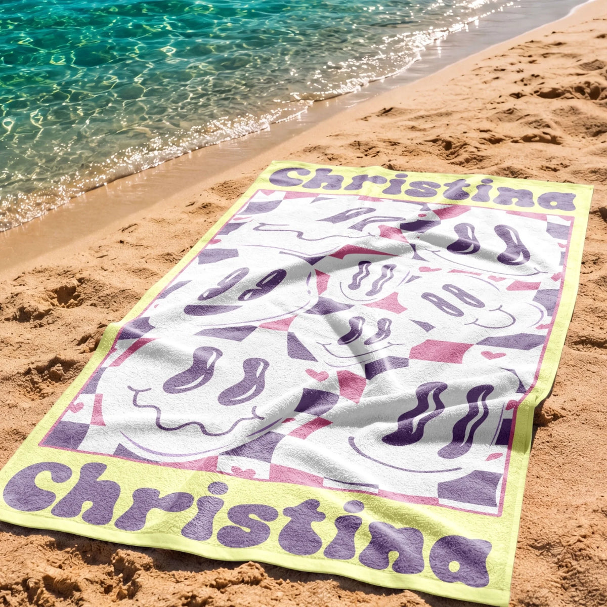 Personalized beach towel with a wavy smiley face theme and the name Christina, laid out on sandy beach shoreline.