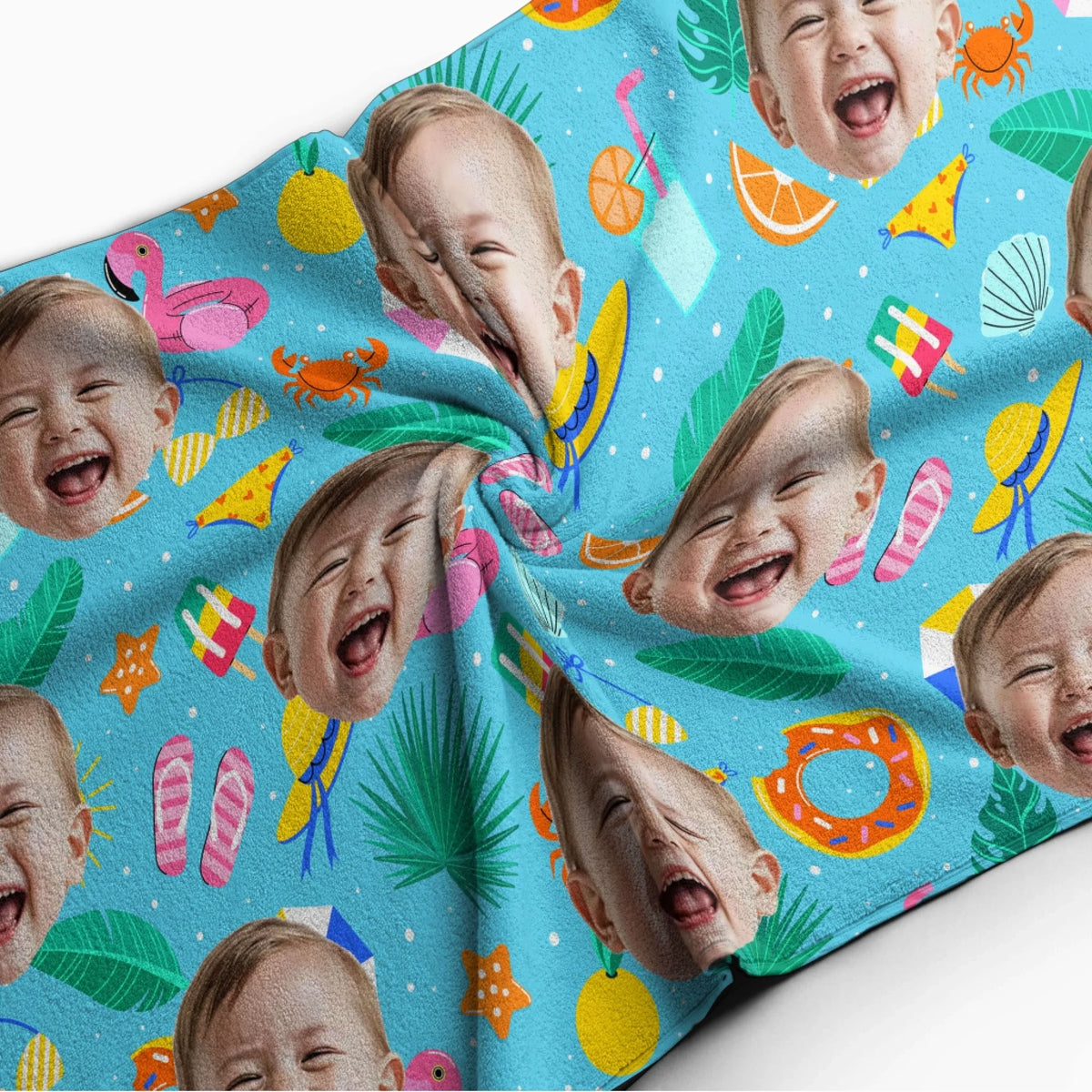 A soft beach towel featuring a repeating pattern of a smiling baby face, pink flamingos, crabs, and tropical fruits.