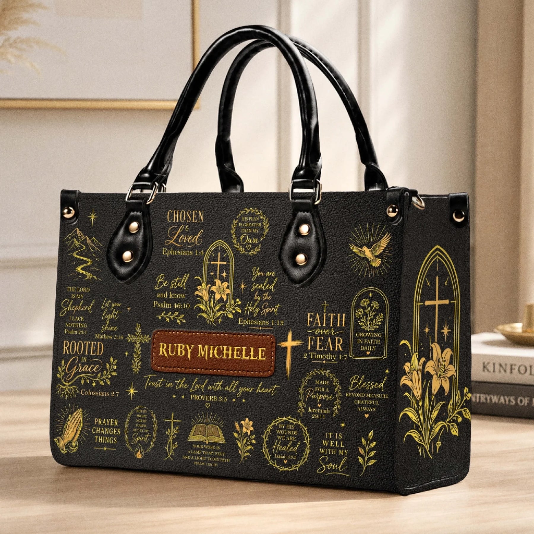Black personalized christian handbag with gold inspirational quotes and floral accents displayed on a wooden table.