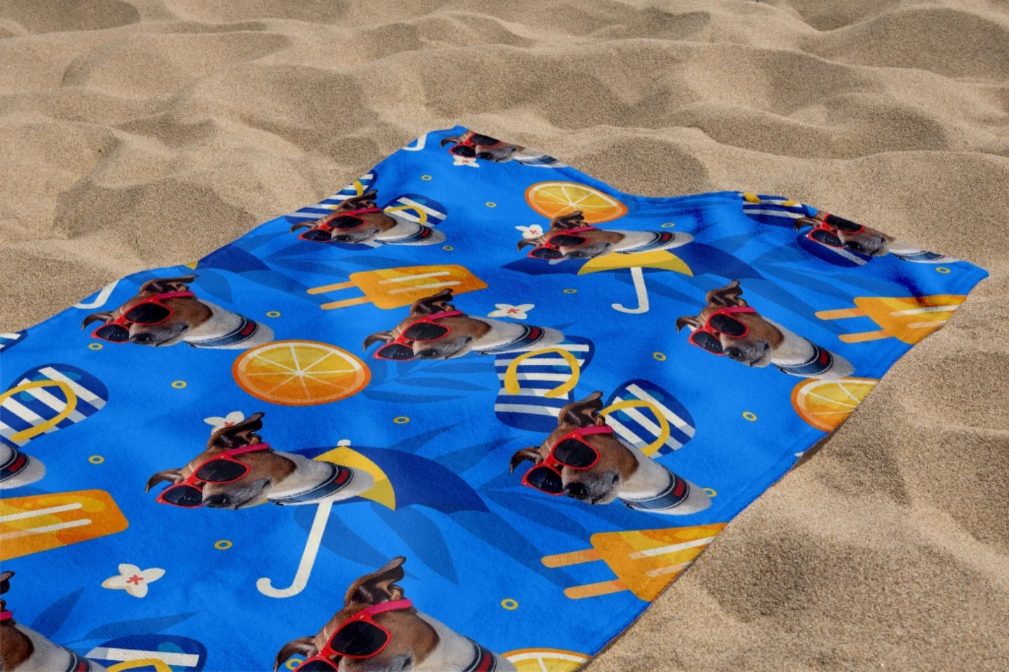 A personalized blue beach towel laid on sand featuring a repeating pattern of a dog face wearing red sunglasses.