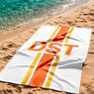 Personalized beach towel featuring orange DST text and bold orange and yellow stripes lying on the sand by the ocean.
