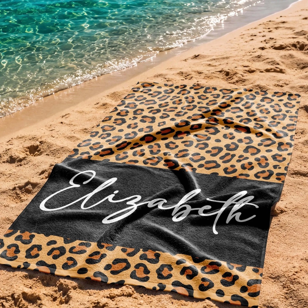 A custom leopard print beach towel featuring the name Elizabeth on a black band, spread out on a sandy beach