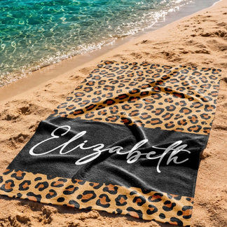 A custom leopard print beach towel featuring the name Elizabeth on a black band, spread out on a sandy beach