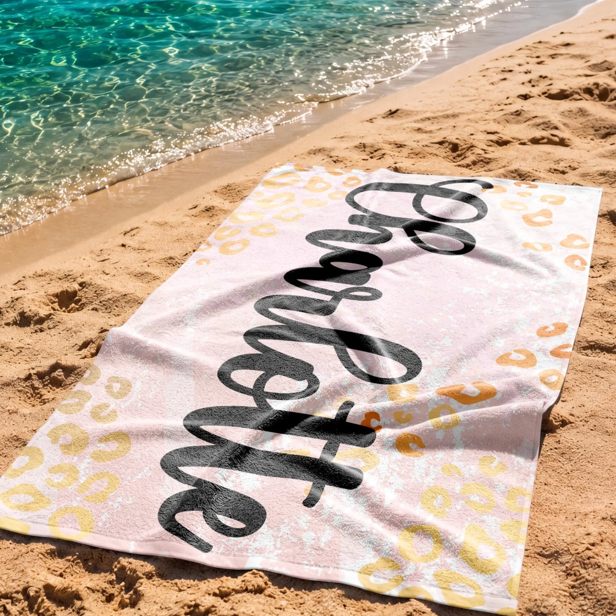 Personalized leopard print beach towel laid on the sand by the water, ideal for a summer beach trip or vacation.