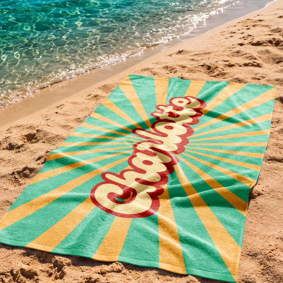 A personalized beach towel with the name Charlotte displayed on a sunburst background, laid on a sandy beach shore.
