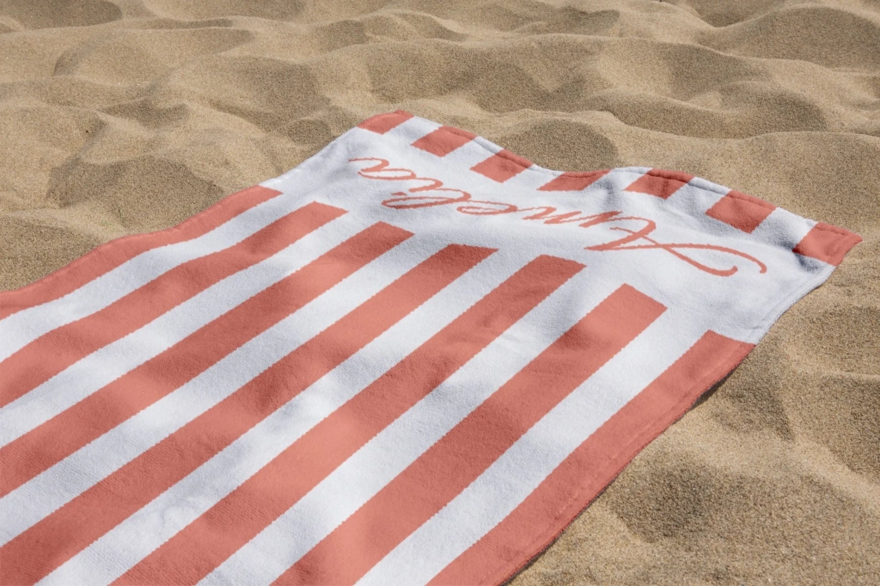 Folded peach and white striped personalized beach towel with Amelia text resting on the sand at a summer beach.