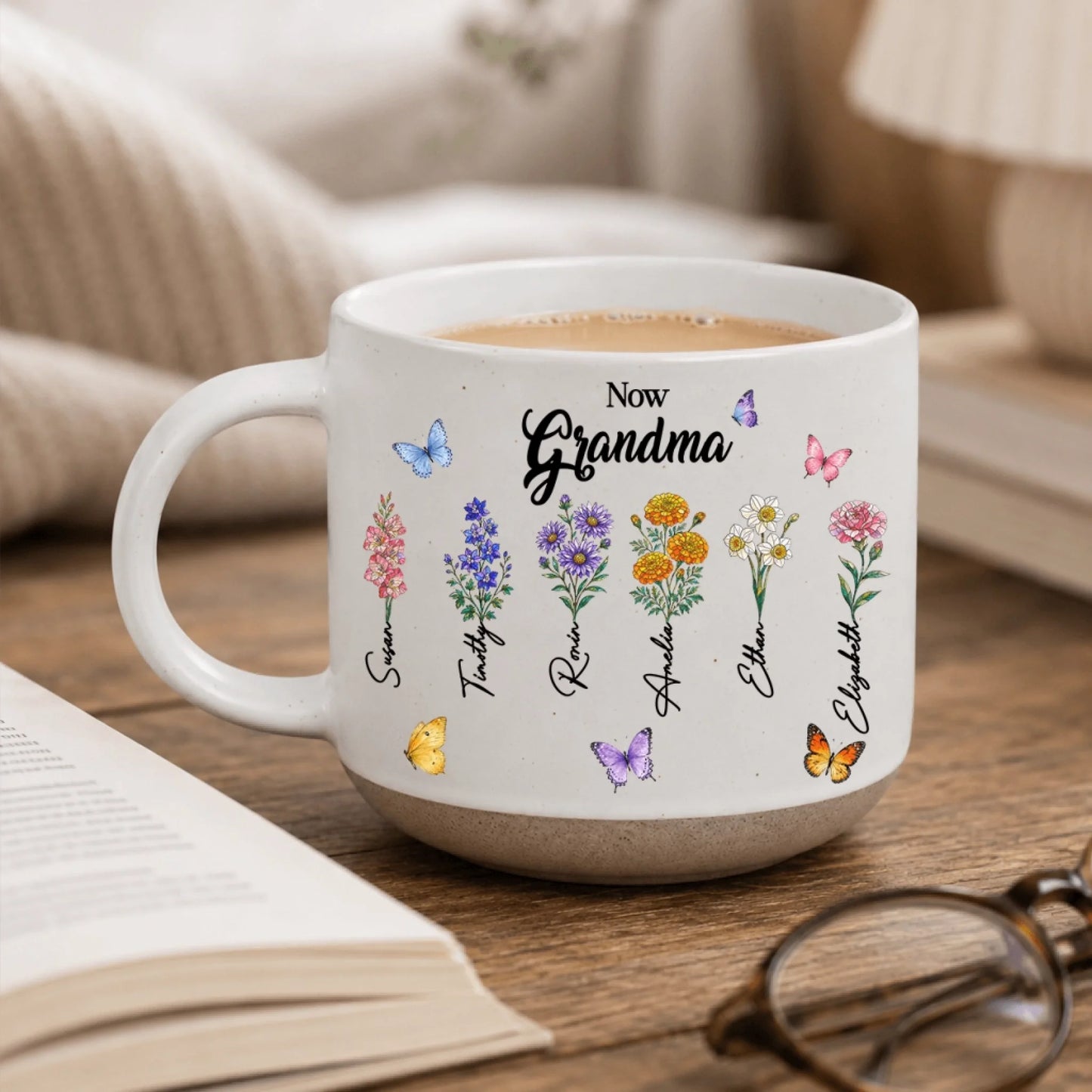 Personalized 'Now Grandma' mug displayed with a book, featuring six floral stems with names and butterflies.
