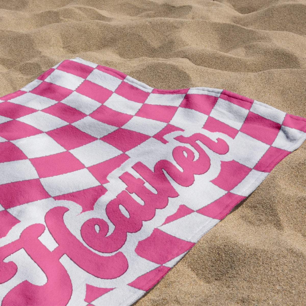 Personalized pink and white checkered beach towel with the custom name Heather lying on a sandy beach shore.