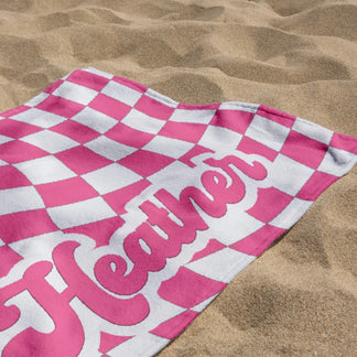 Personalized pink and white checkered beach towel with the custom name Heather lying on a sandy beach shore.