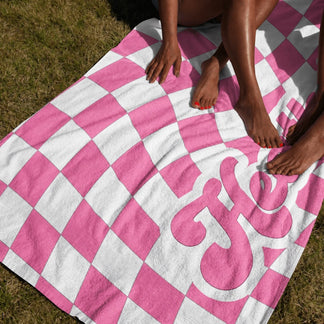 A woman sits on a personalized pink and white checkerboard patterned beach towel with the name Heather printed on it.