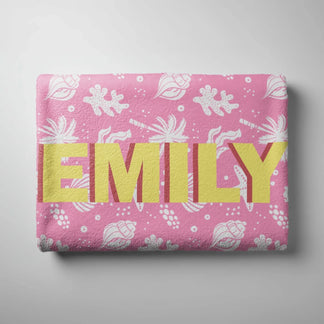 Personalized pink beach towel with Emily in yellow text surrounded by a white tropical seashell and palm design.