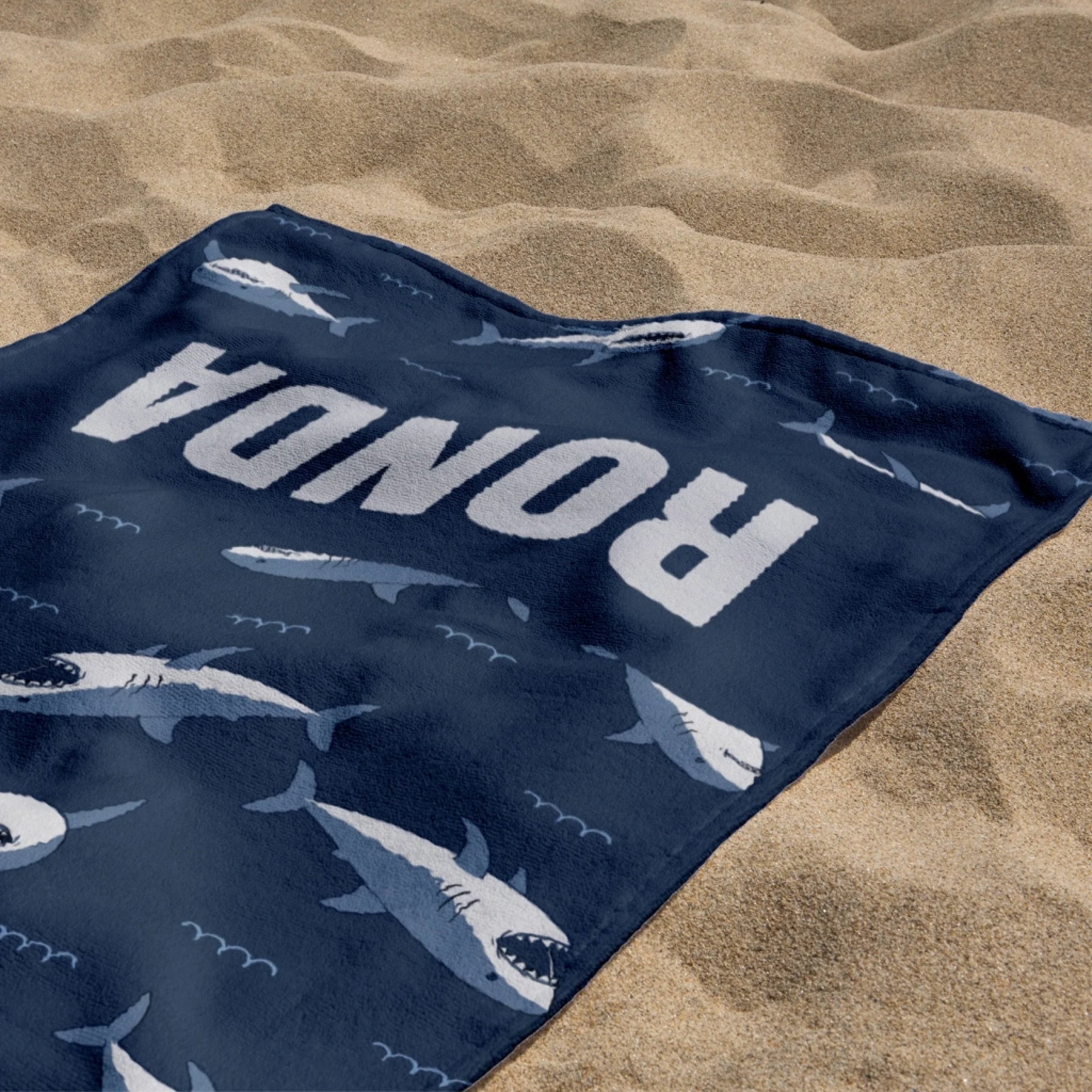 A dark blue personalized beach towel with a shark print and the name Ronda spread out on warm beach sand.