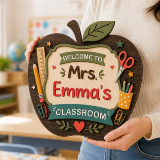 Person holding a personalized wooden apple sign that reads Mrs. Emma's Classroom in a classroom setting.