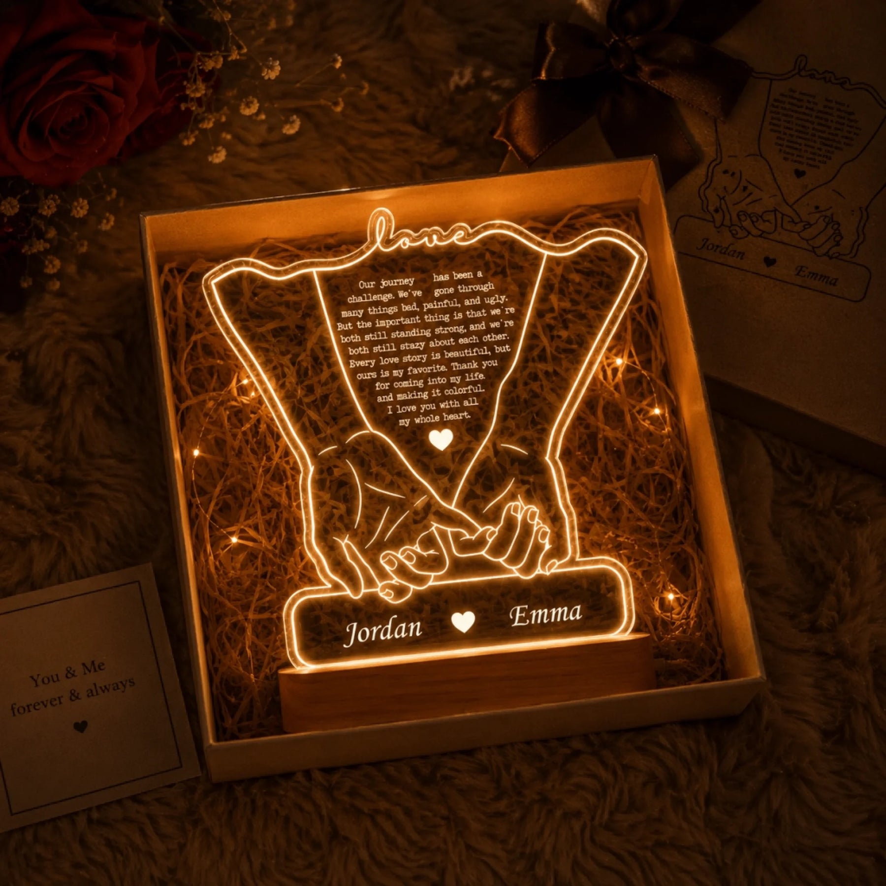 An illuminated acrylic lamp in a gift box, showing two hands holding. It features a heartfelt message and names on a wooden base, surrounded by roses and a small card.