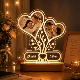 A personalized acrylic night light displaying two heart-shaped photos of a couple, a tree motif, and the names Daniel and Olivia, glowing warmly on a table in a romantic setting.