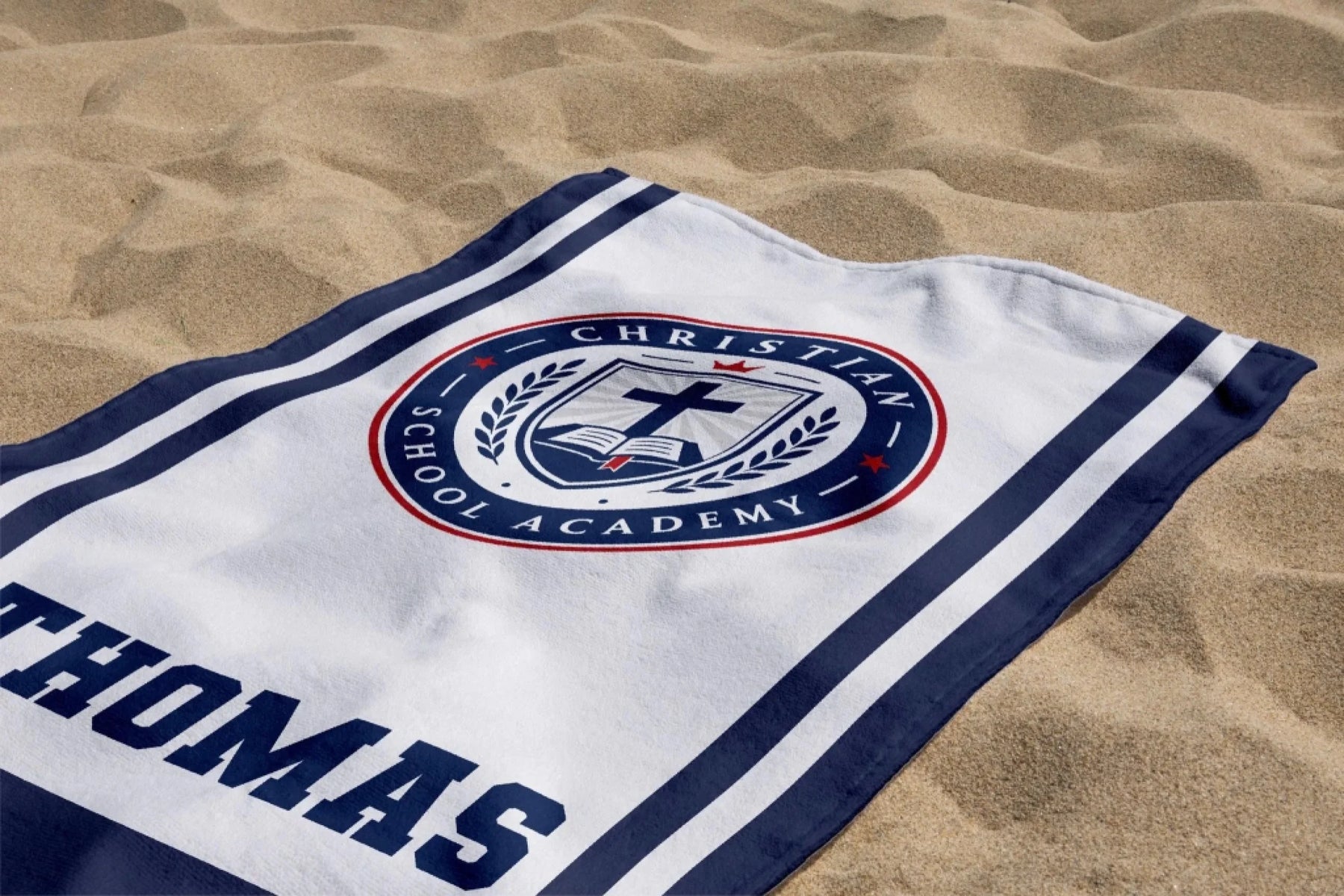 Personalized school spirit beach towel with Christian School Academy emblem and Thomas name laid on the sand.