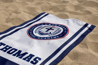 Personalized school spirit beach towel with Christian School Academy emblem and Thomas name laid on the sand.