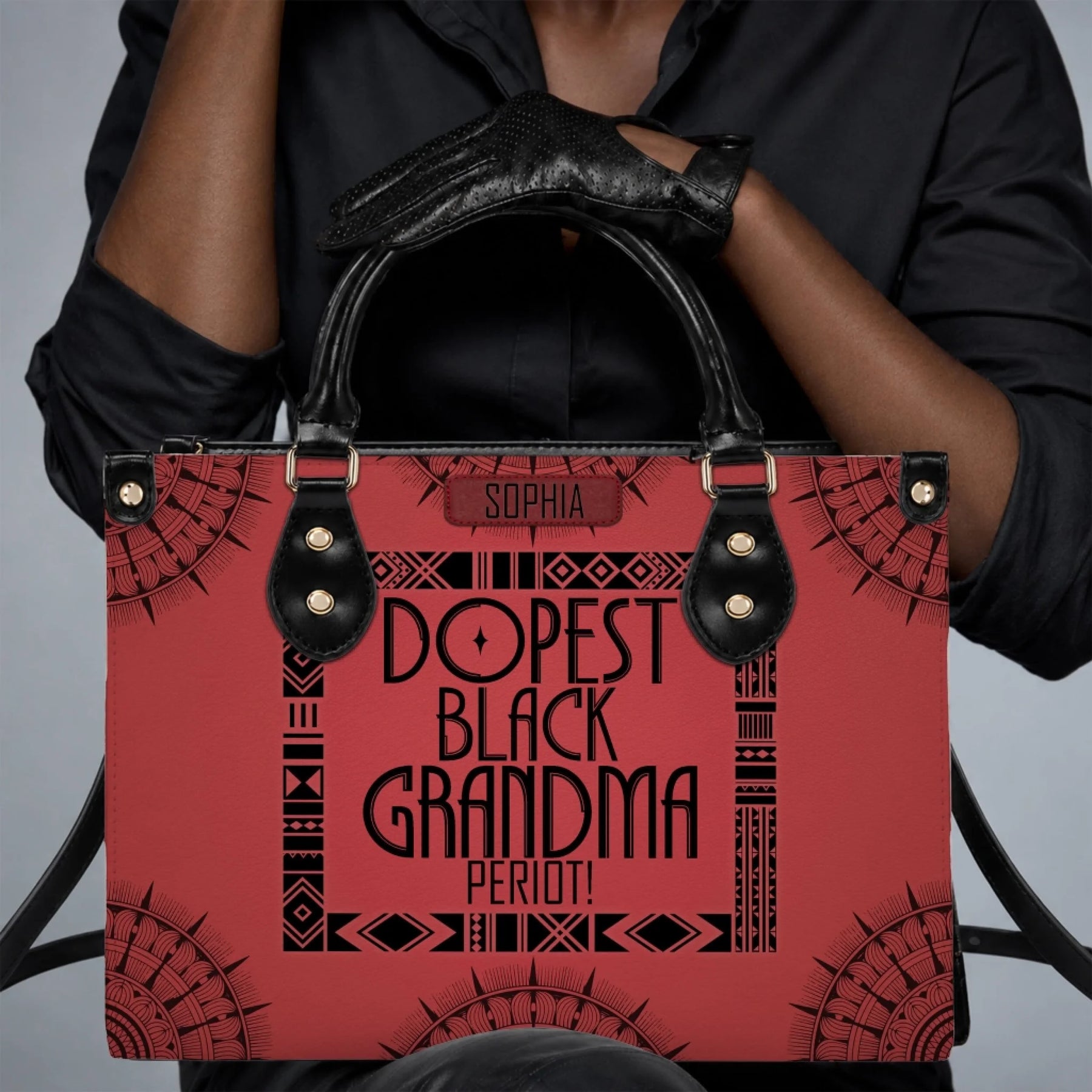 Close up of a person wearing black gloves holding a personalized red handbag labeled Dopest Black Grandma Period.