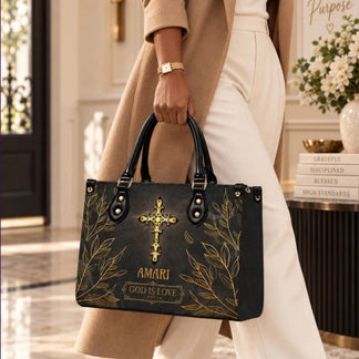 A woman wearing a tan coat holds a black tote bag with a golden cross emblem and the customized text Amari God is Love.