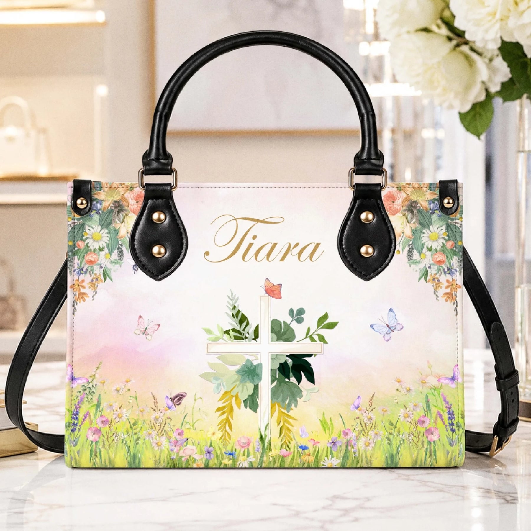 A stylish personalized handbag featuring a watercolor cross and butterfly design with the custom name Tiara on a marble table.