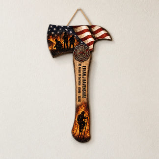 A decorative axe plaque with an American flag, firefighter silhouettes, an emblem, a name, and service dates hangs on a white wall.