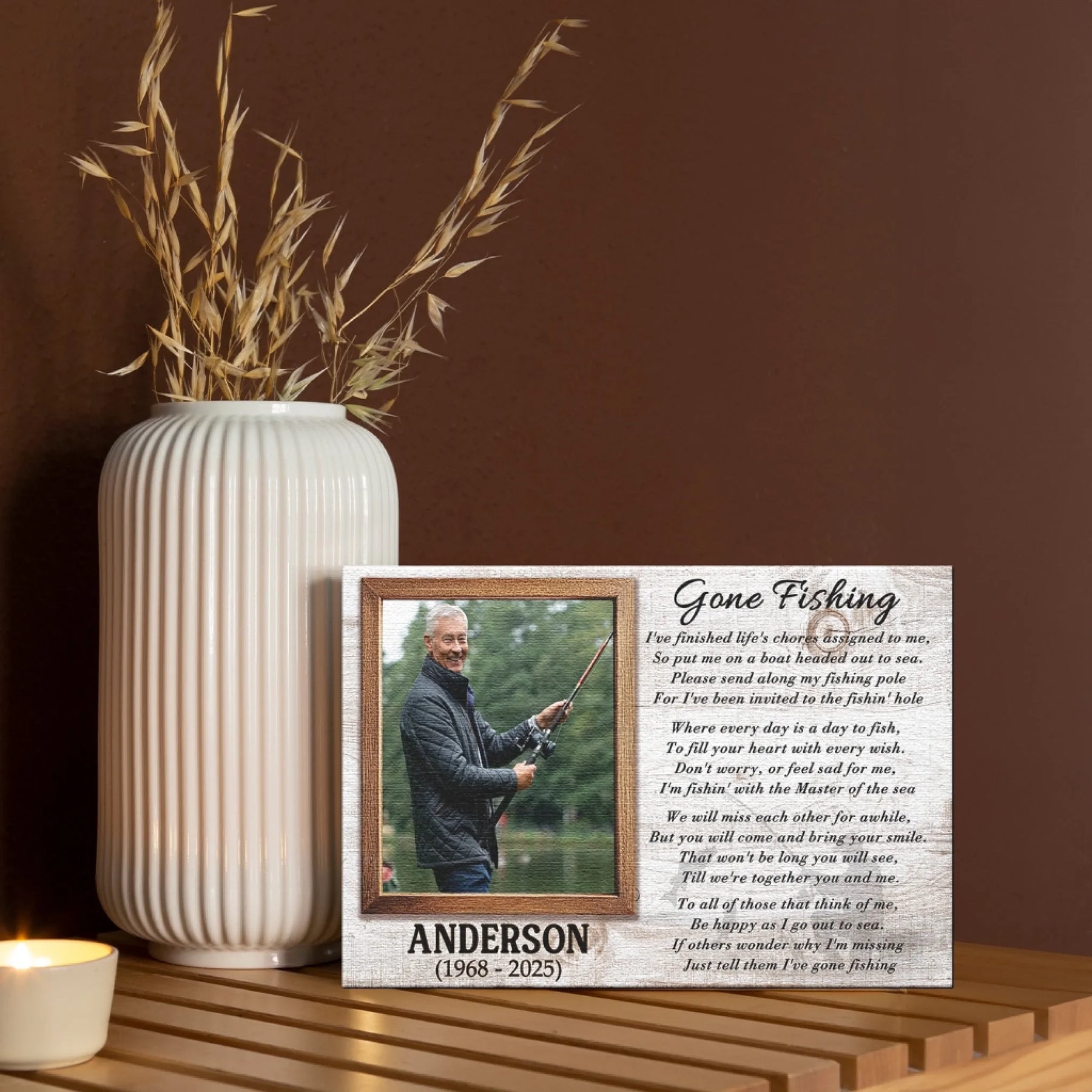 A rectangular canvas print with a fishing photo, poem, and name, displayed on a wooden shelf next to a white vase and candle.