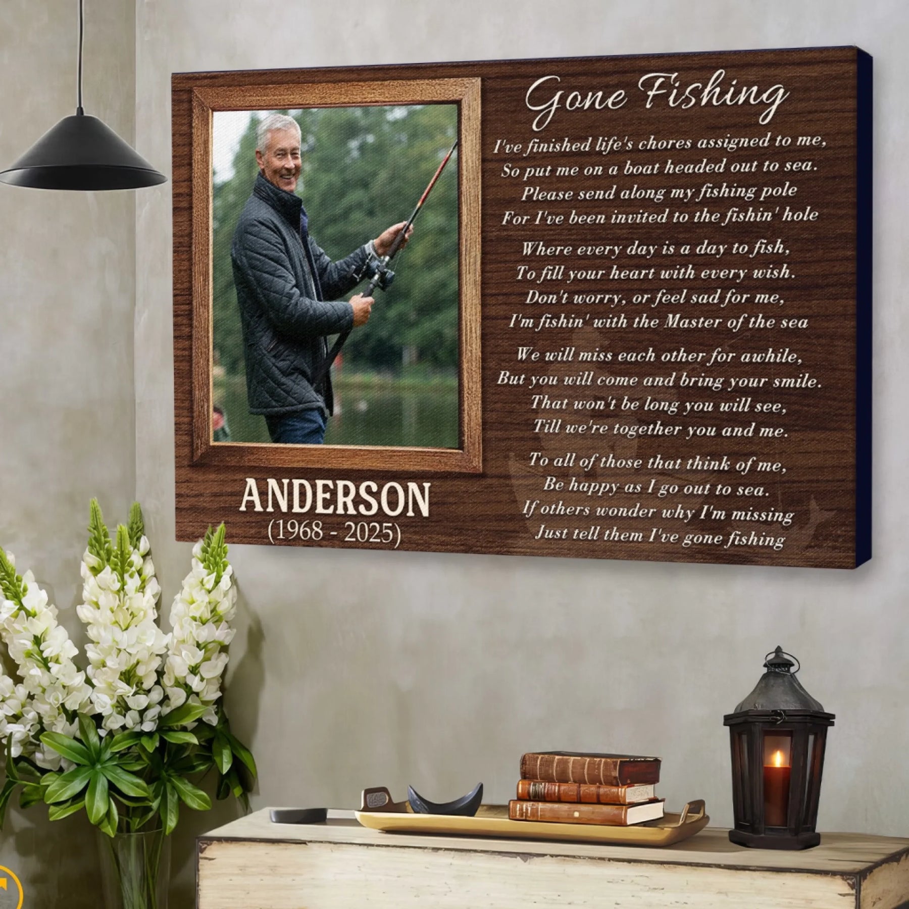 A wooden plaque featuring a fishing photo and poem hangs on a textured wall, above a chest with white flowers, books, and a candle lantern.