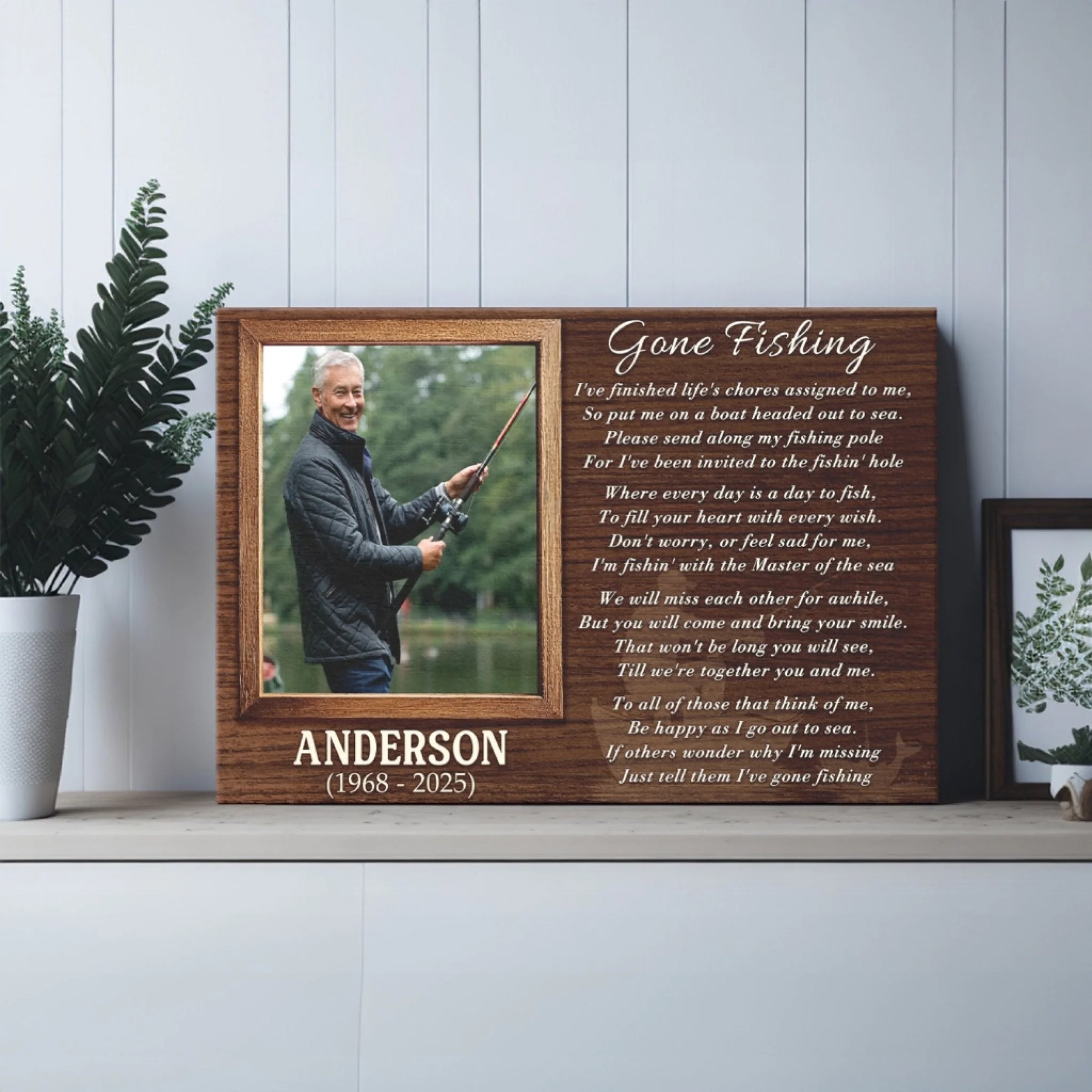 A wooden plaque with a fishing photo and poem rests on a shelf in a room with a green plant and a framed botanical print on the right.