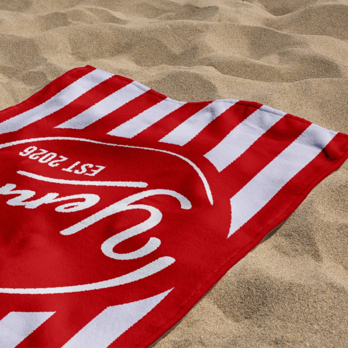 A folded red and white striped beach towel with custom Yenna and EST 2026 text resting on the sandy beach.