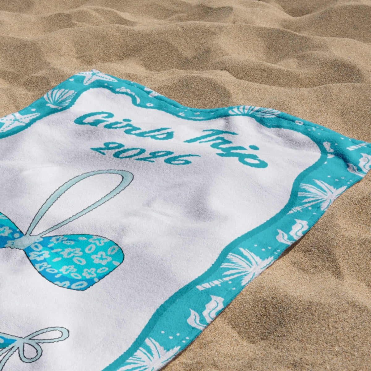 A personalized beach towel for a 2026 girls trip laid out on sand with a bikini design and teal coastal border.