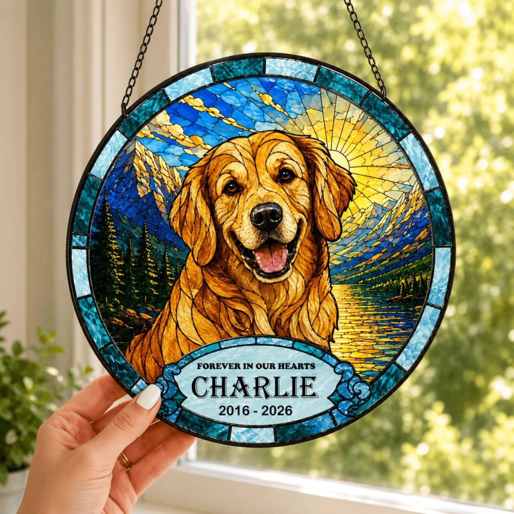 A hand holds a round stained-glass style suncatcher depicting a smiling golden dog in a mountain and lake landscape. A banner shows memorial text.