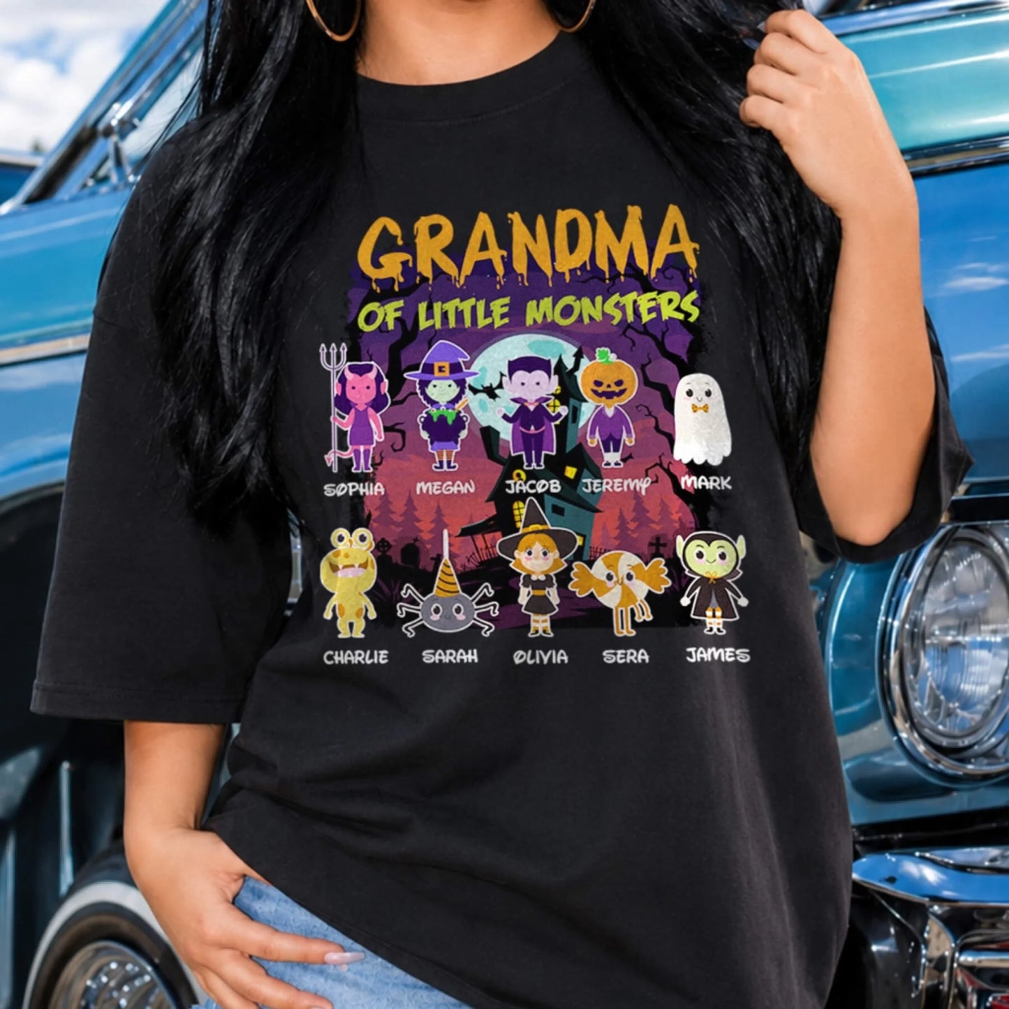 A woman wears a black personalized Halloween t-shirt featuring a Grandma of Little Monsters design with custom names.