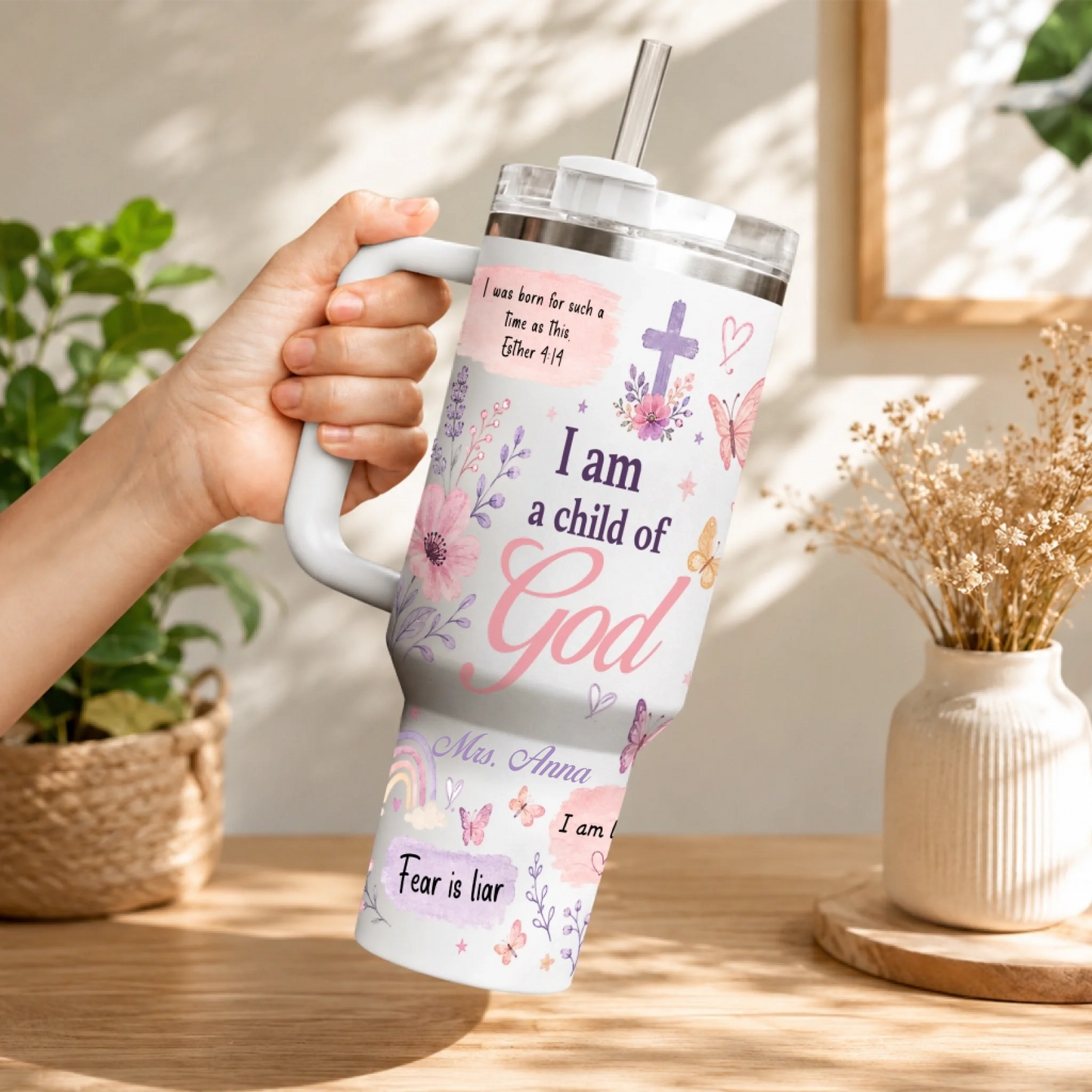 A woman's hand holding a personalized floral Christian tumbler with straw, featuring 'I am a child of God' and butterflies.