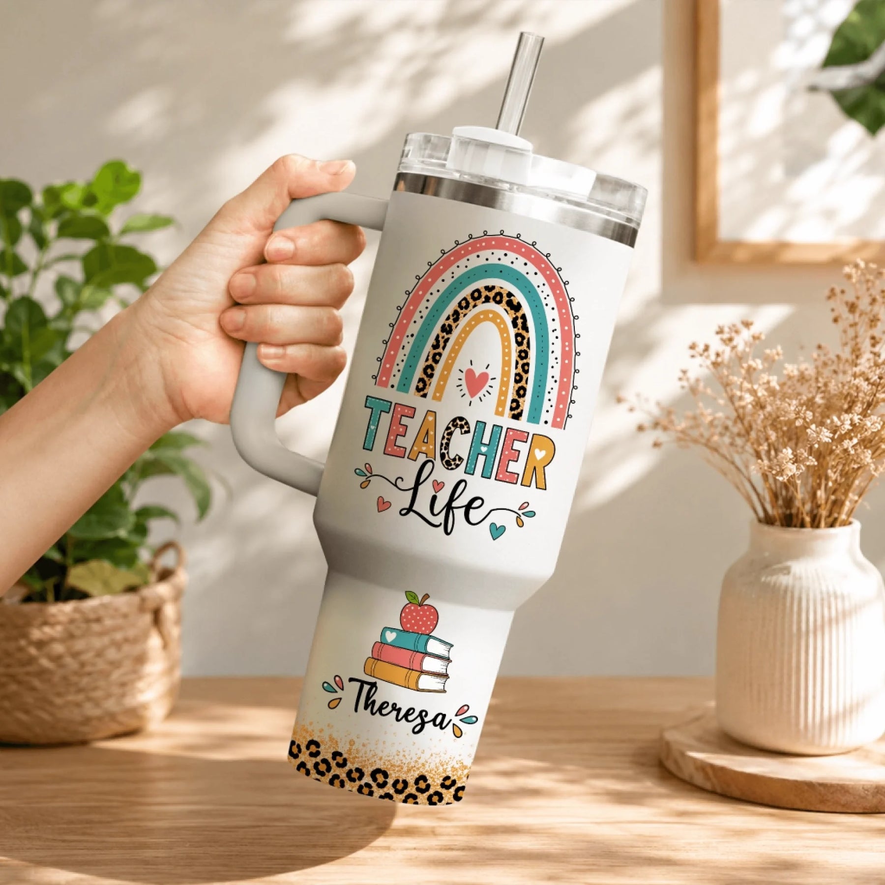 Hand holding a white stainless steel teacher life tumbler with custom name Theresa, rainbow, and leopard print accents.