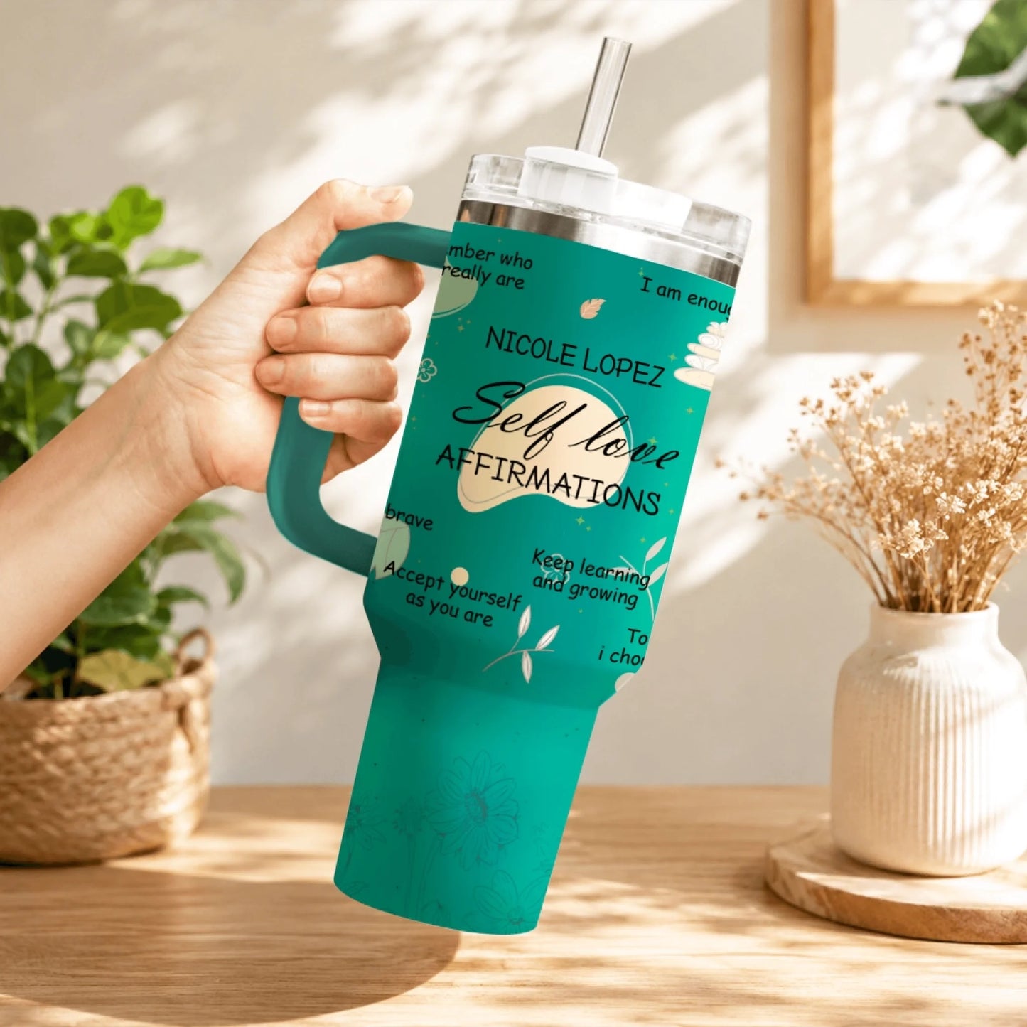 A hand holding a teal stainless steel tumbler decorated with inspiring self-love quotes and flower designs.
