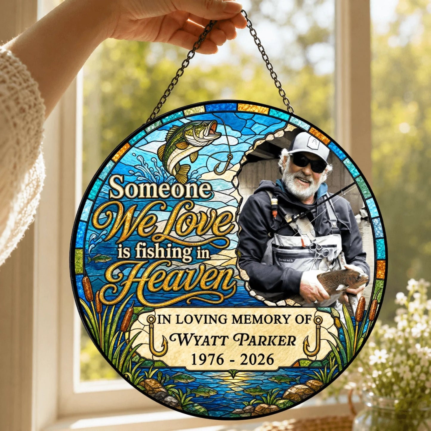 A hand holding a round stained-glass memorial ornament with a fishing theme, hanging in front of a bright window.