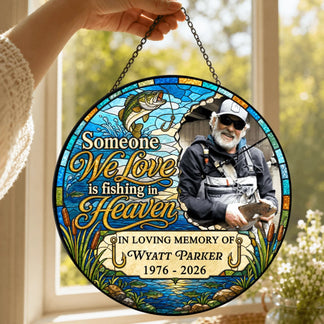 A hand holding a round stained-glass memorial ornament with a fishing theme, hanging in front of a bright window.
