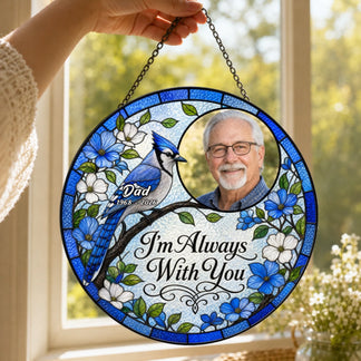 A person holding a round stained glass memorial ornament with a blue jay, flowers, and a photo of a man.