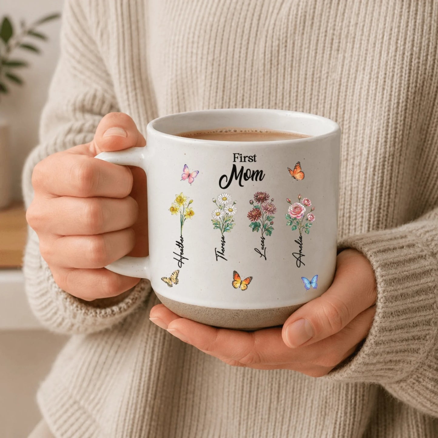 A person wearing a beige sweater holding a personalized white First Mom ceramic mug with colorful flower art.
