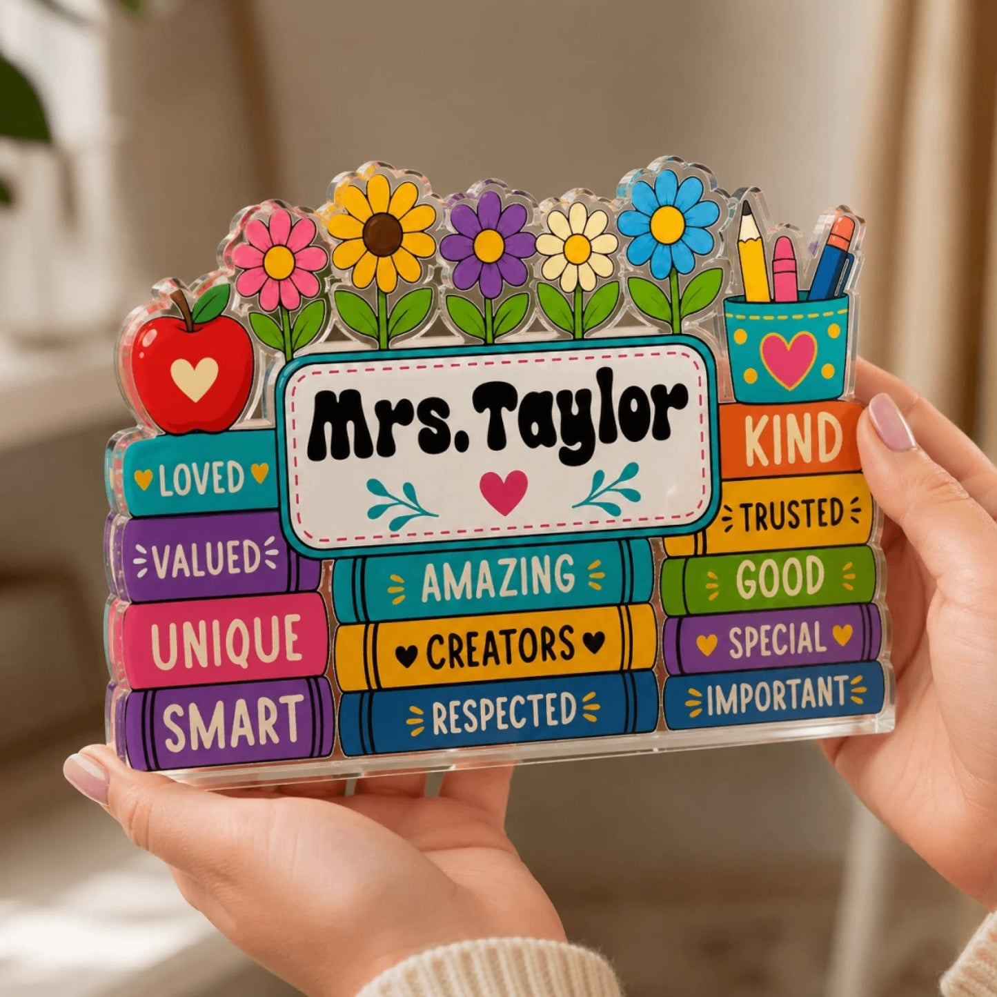 Hands holding a custom acrylic desk plaque for a teacher labeled Mrs. Taylor with colorful book and flower design.