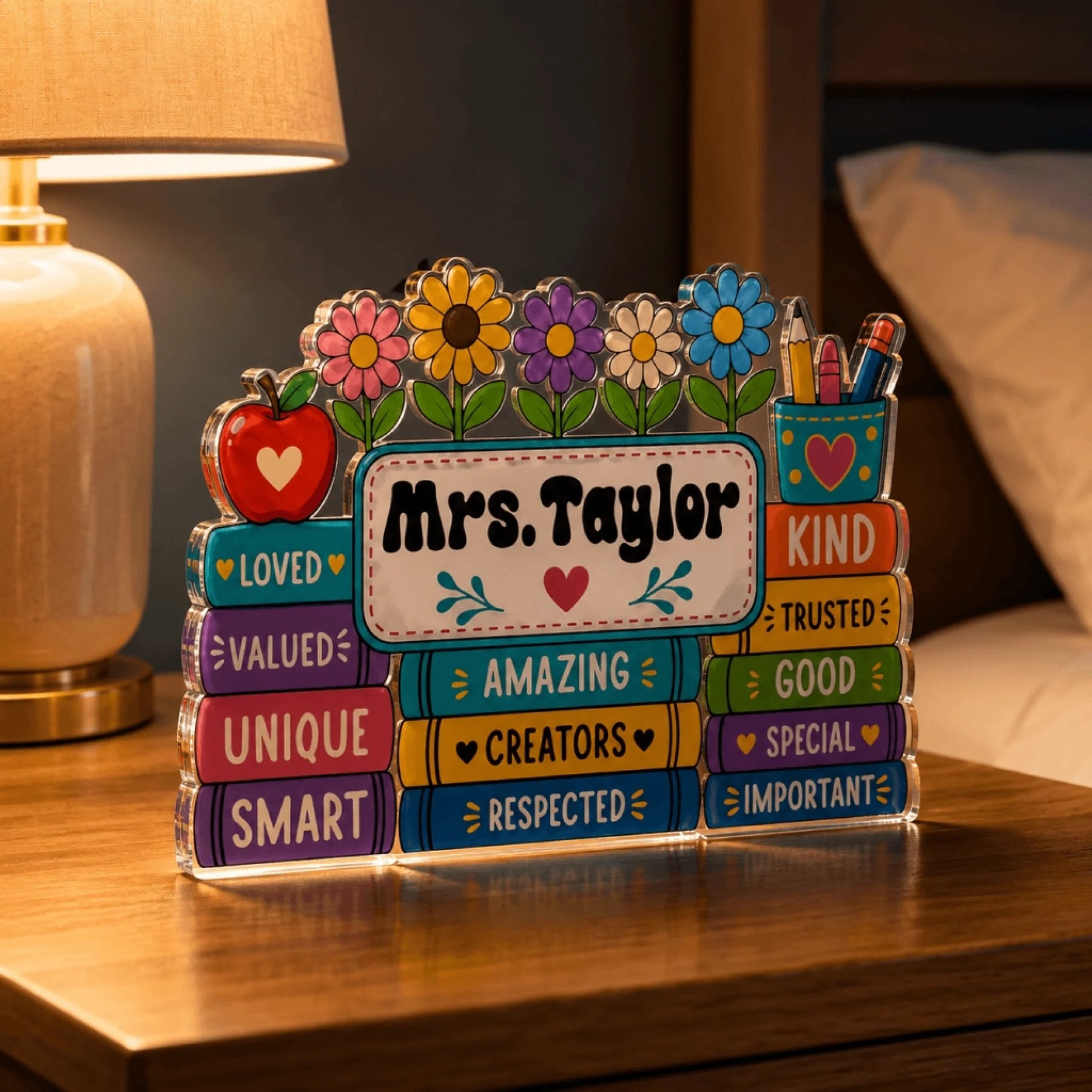 Decorative acrylic teacher desk sign titled Mrs. Taylor displayed on a wooden desk near a warm lamp.